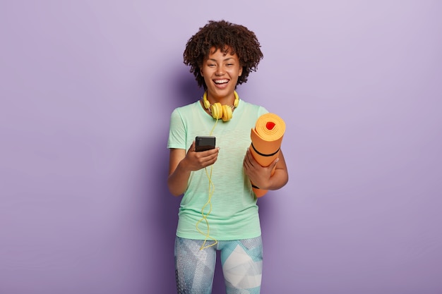 Concepto de entrenamiento y fitness. Alegre mujer de piel oscura tiene teléfono móvil conectado a auriculares