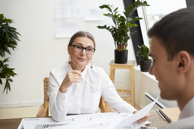 Concepto de equipo, cooperación y creatividad de ensueño. Hermosa alegre arquitecta mujer madura vistiendo blusa blanca y gafas alabando a su talentoso y ambicioso colega masculino joven, revisando sus dibujos