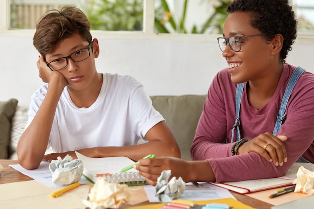 Foto gratuita concepto de escuela y educación. una maestra con experiencia y complacida ayuda al joven a ponerse al día con el grupo, explica la regla gramatical y toma notas en la libreta. adolescente siente apatía porque no quiere estudiar