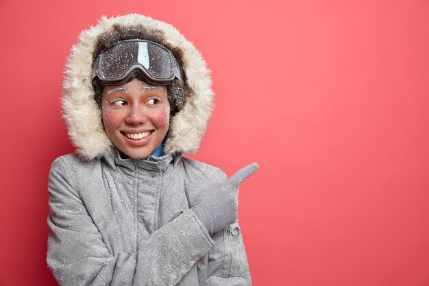 Concepto de esquí y diversión de invierno. La mujer afroamericana de piel oscura complacida en ropa de abrigo apunta hacia el espacio en blanco que muestra la dirección al lugar del resort disfruta de la temporada de diciembre.