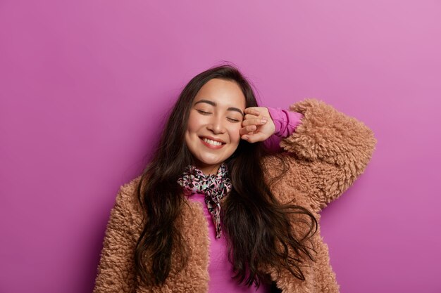 Concepto de estado de ánimo feliz. Mujer joven soñadora relajada inclina la cabeza, sonríe suavemente, vestida con ropa de abrigo, mantiene los ojos cerrados, aislado sobre el espacio lila