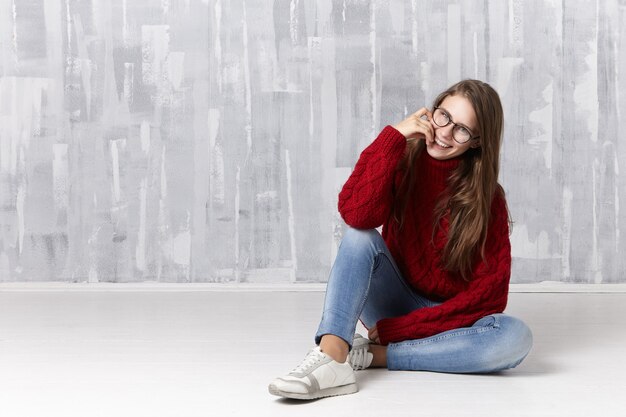 Concepto de estilo, belleza, moda, ropa y gafas. Adolescente de aspecto moderno con cabello suelto sentada en el piso, sonriendo juguetonamente, tocándose los labios, con gafas, suéter, jeans y zapatillas de deporte