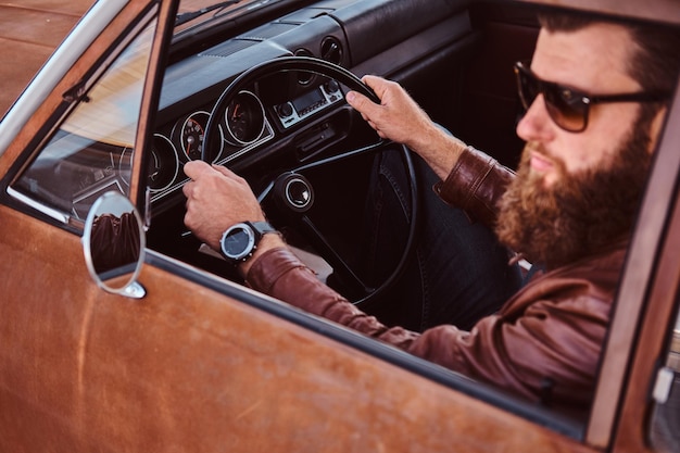 Concepto de estilo campestre. Hombre barbudo con gafas de sol vestido con chaqueta de cuero marrón conduciendo un camión retro.