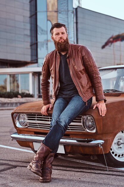 Concepto de estilo campestre. Hombre barbudo vestido con chaqueta de cuero marrón y botas apoyado en un auto retro sintonizado en el estacionamiento de la ciudad cerca de un rascacielos.