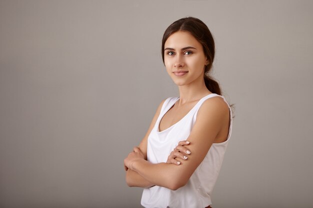 Concepto de estilo, confianza y personas. Tiro de cintura para arriba de mujer joven atractiva en casual blanco superior cruzando los brazos sobre su pecho de buen humor, con sonrisa positiva y segura