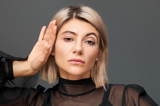 Concepto de estilo, glamour, cosmética y belleza. Retrato de mujer joven impresionante en blusa negra transparente posando aislado conmovedor rostro, con piercing facial, corte de pelo corto teñido y maquillaje de moda