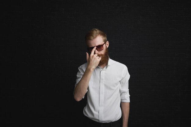 Concepto de estilo y moda. Apuesto joven de moda con barba pelirroja posando en ropa formal, ajustando elegantes gafas de sol, con expresión de confianza