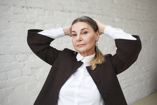 Foto gratuita concepto de estilo y moda. disparo aislado horizontal de seria exitosa empresaria caucásica de 55 años con cabello recogido cogidos de la mano detrás de la cabeza, mirando con mirada de confianza