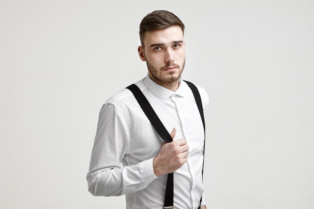 Concepto de estilo, moda y elegancia. Imagen del joven gerente masculino barbudo caucásico de moda con camisa blanca mirando a la cámara con expresión seria y segura, tirando de sus tirantes