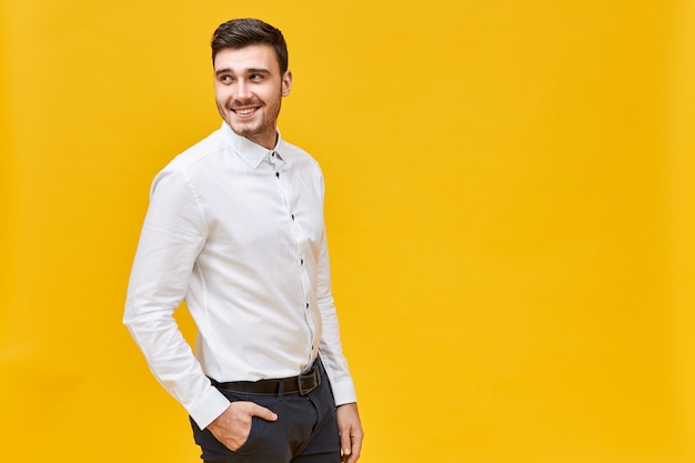 Concepto de estilo, moda y ropa de hombre. Apuesto joven empresario positivo posando aislado con la mano en el bolsillo de elegantes jeans negros, mirando hacia atrás, con expresión facial alegre