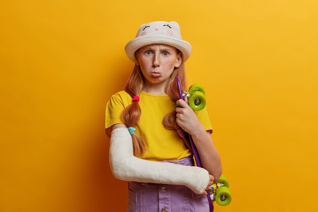 Concepto de estilo de vida activo, infancia y lesiones. Adorable niña pecosa posa con penny board y brazo roto, se lesionó durante el skate y haciendo trucos peligrosos. Pasatiempo de los adolescentes