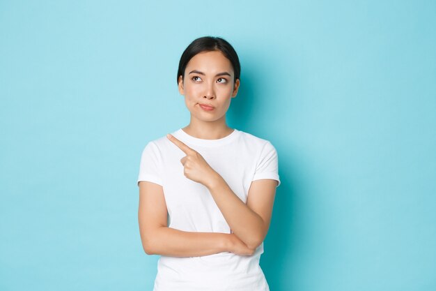 Concepto de estilo de vida, belleza y compras. Mujer asiática escéptica y poco divertida en camiseta blanca apuntando a la esquina superior izquierda y con una sonrisa disgustada, juzgando algo, de pie en la pared azul.