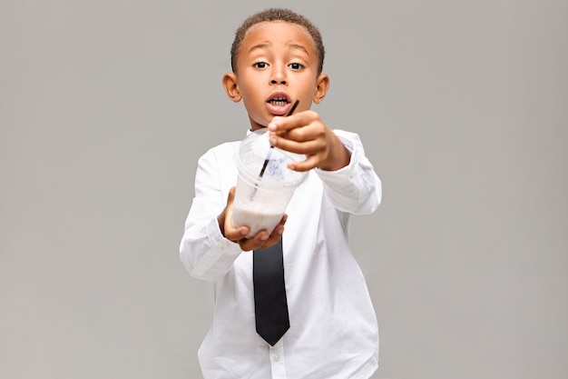 Concepto de estilo de vida, comida, bebida e infancia saludable. Imagen de un colegial afroamericano emocionado emocionalmente en uniforme sosteniendo un frasco de plástico con batido de frutas, ofreciéndole tener algunos