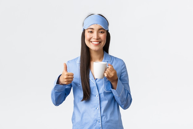 Concepto de estilo de vida, desayuno y personas de la mañana. Alegre y sonriente chica bastante asiática con máscara para dormir y pijama mostrando el pulgar hacia arriba mientras toma café después de despertarse, sintiéndose lleno de energía.