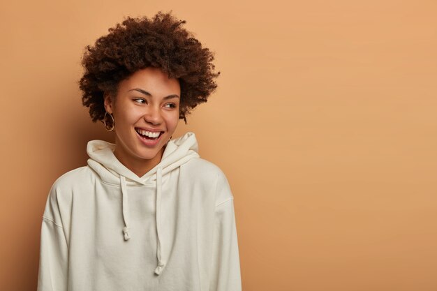 Concepto de estilo de vida y emociones. Feliz mujer rizada de piel oscura encantada viste sudadera blanca, se ríe y se divierte, mira a un lado