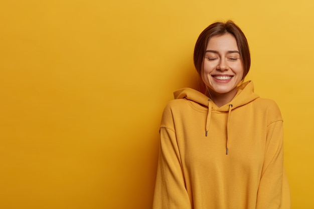 Concepto de estilo de vida y emociones. La mujer alegre cierra los ojos y se ríe, se ríe y se divierte, usa una sudadera, muestra los dientes blancos