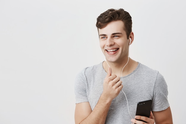 Concepto de estilo de vida y entretenimiento. Apuesto hombre guapo con cabello oscuro y ojos azules se ve feliz mientras escucha canciones agradables en los auriculares, recuerda momentos agradables de la vida, mira a un lado.