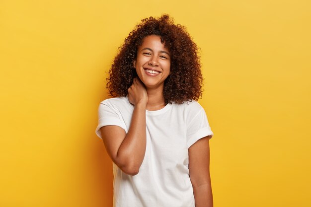 Concepto de estilo de vida feliz. Agradable y divertida mujer afro se siente afortunada y satisfecha, se ríe feliz, tiene dientes blancos con un pequeño espacio, disfruta de un día libre increíble, se para contra la pared amarilla