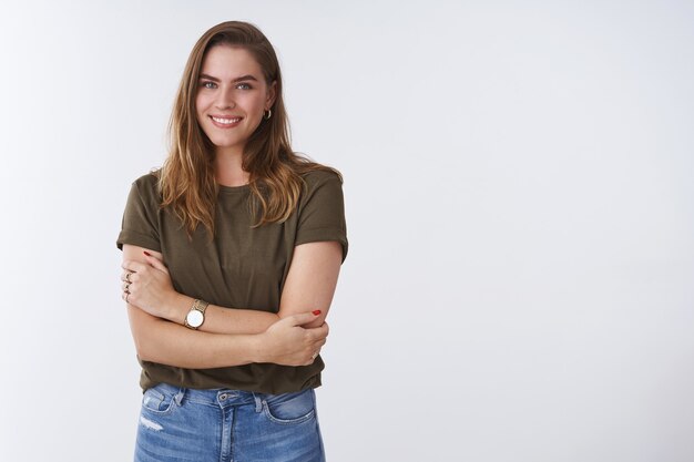 Concepto de estilo de vida de personas de bienestar. Encantadora mujer joven saliente tierna vistiendo camiseta verde oliva pecho de brazos cruzados sonriendo lindo coqueto expresando positividad actitud alegre, fondo blanco.