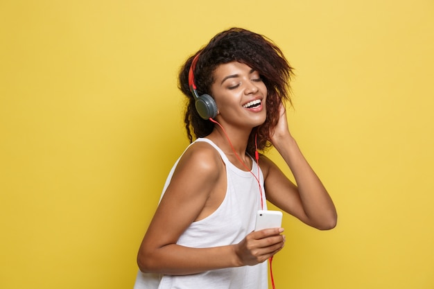 Concepto de estilo de vida - Retrato de hermosa mujer afroamericana alegre escuchar música en el teléfono móvil. Fondo amarillo pastel del estudio. Espacio De La Copia.