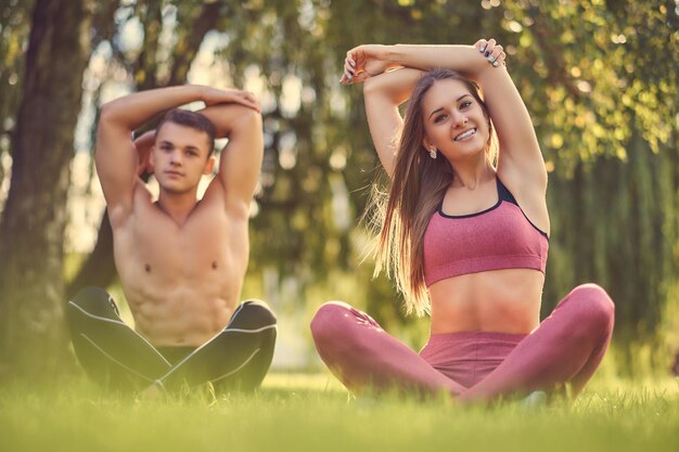 Concepto de estilo de vida saludable. Una feliz pareja joven de fitness estirando las manos mientras se sienta en una pose de loto sobre un césped verde.