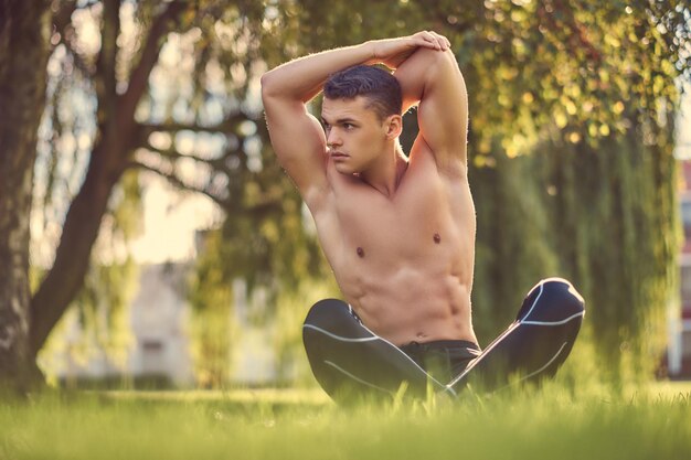 Concepto de estilo de vida saludable. Un joven sin camisa estirando las manos mientras se sienta en una pose de loto sobre hierba verde.