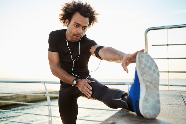 Foto gratuita concepto de estilo de vida saludable. joven corredor masculino de piel negra con cuerpo atlético que se extiende en el muelle, colocando su pierna en la plataforma, calentando