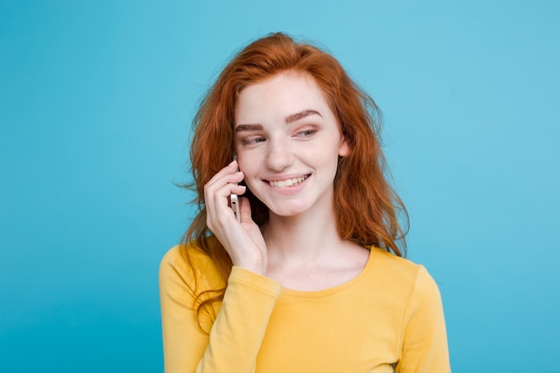 Concepto de estilo de vida y tecnología - Retrato de alegre alegre pelo rojo jengibre chica alegre y emocionante hablando con su amigo por teléfono móvil. Aislado en el fondo azul Pastel. Copie el espacio.