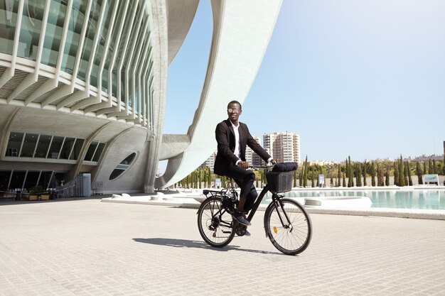 Concepto de estilo de vida urbano, ecología y transporte. Joven empresario afroamericano moderno, ecológico y moderno que usa tonos redondos de moda y un traje formal de ciclismo para trabajar en bicicleta