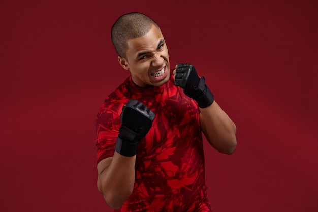 Foto gratuita concepto de éxito, determinación, desafío y competencia. imagen del joven luchador afroamericano agresivo furioso con guantes de entrenamiento negros de boxeo en el estudio, rugiendo, preparándose para la pelea