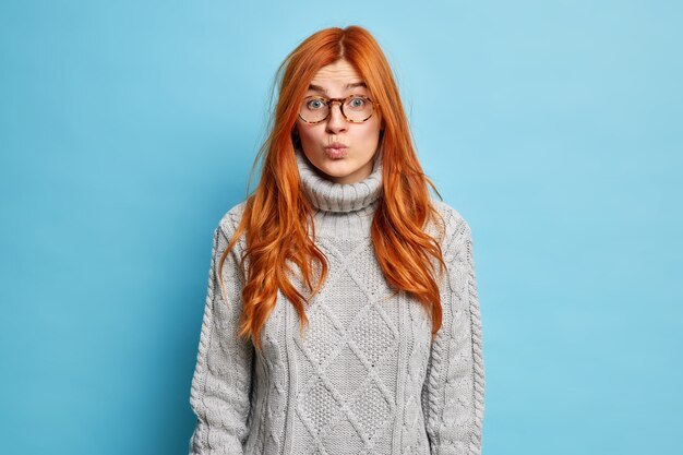 Concepto de expresiones faciales. Bonita mujer europea pelirroja mantiene los labios doblados y luce sorprendentemente viste suéter de punto.