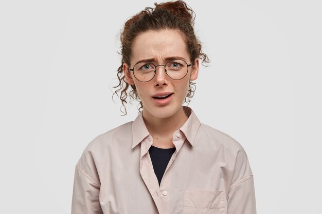 Concepto de expresiones faciales negativas. Infeliz mujer pecosa indignada con cabello rizado, frunce el ceño con disgusto, viste camisa beige de gran tamaño, escucha algo desagradable del interlocutor