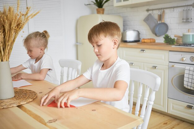 Concepto de familia e infancia. Retrato de dos hermanos varones en edad escolar sentados juntos a la mesa en la cocina: niño rubio haciendo la tarea mientras su hermano mayor hace origami en primer plano