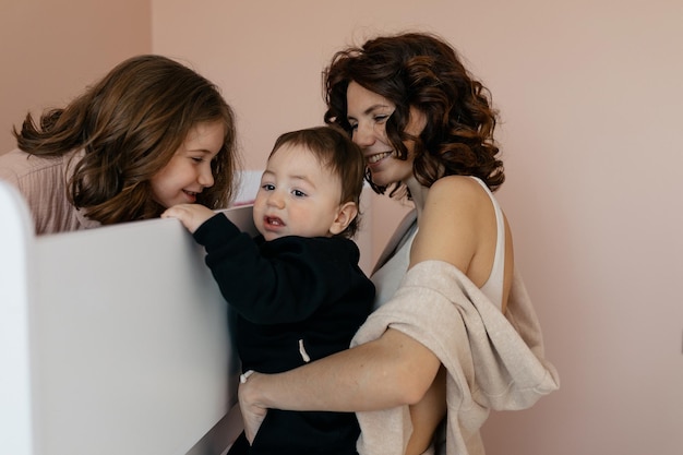 Foto gratuita concepto de familia feliz mujer joven con peinado ondulado en ropa de casa está sosteniendo a su pequeño hijo y besando a su pequeña hija verdaderas emociones felices familia feliz en casa