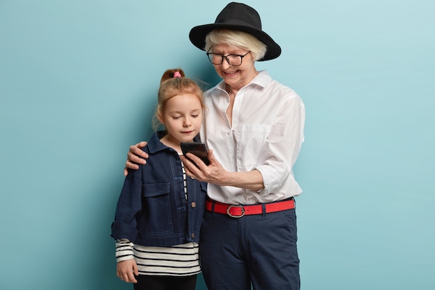 Concepto de familia, generación y tecnología. Feliz abuela de moda sostiene el teléfono inteligente, abraza a la nieta, mira videos juntos, se divierten juntos en casa, posan sobre la pared azul