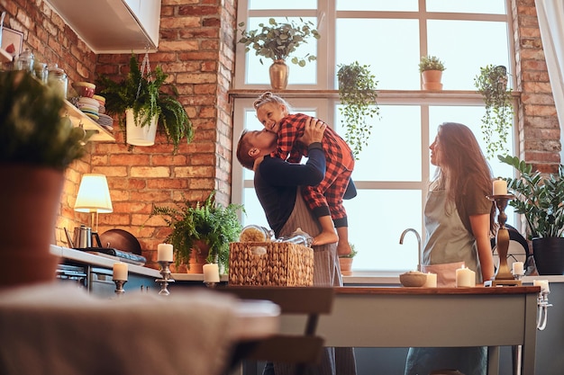 concepto de familia. Papá feliz sostiene a su niña en las manos mientras mamá está parada cerca de la cocina estilo loft por la mañana.