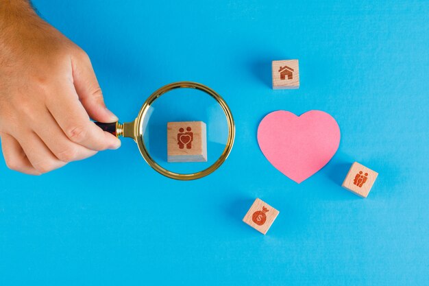 Concepto de familia con papel cortado corazón en mesa azul plano lay. Mano que sostiene la lupa sobre el cubo de madera.