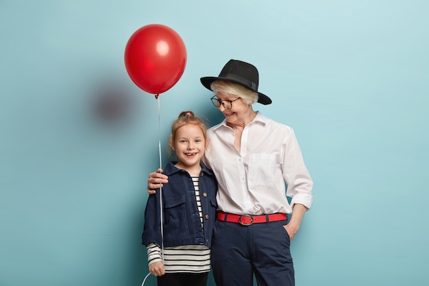 Concepto de familia y vacaciones. cuidando a la abuela de pelo gris con ropa de moda, abraza a la pequeña nieta, celebran juntos el día del niño, se para sobre la pared azul con un globo de decoración