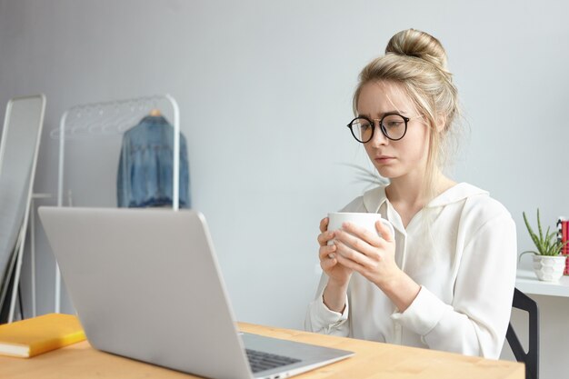 Concepto de fecha límite y exceso de trabajo. Joven mujer caucásica independiente frustrada en elegantes gafas bebiendo otra taza de café mientras trabaja en un proyecto urgente, sentada frente a una computadora portátil abierta