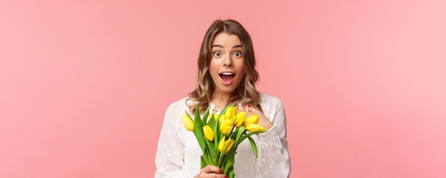Concepto de felicidad y celebración de primavera El primer plano de una joven rubia sorprendida y asombrada con vestido blanco recibe tulipanes amarillos y se señala a sí misma con incredulidad y asombro.