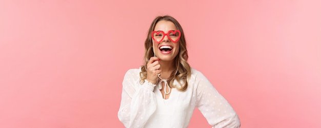 Concepto de felicidad y celebración de primavera Retrato de mujer caucásica hermosa divertida y despreocupada con vestido blanco de pelo rubio con máscara de gafas en forma de corazón y riendo