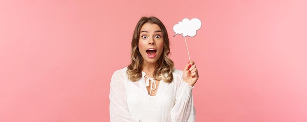 Concepto de felicidad y celebración de primavera Retrato de primer plano de una chica rubia emocionada sorprendida que parece asombrada sosteniendo un palo de comentarios en la nube cerca de la cabeza y jadeando asombrado tiene una idea de fondo rosa