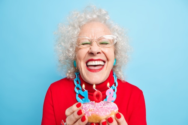 Concepto de feliz cumpleaños. Dama mayor encantada sonríe ampliamente tiene dientes blancos perfectos que van a soplar velas en donas glaseadas estar bien vestidas celebra el 102 ° cumpleaños