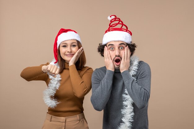 Concepto festivo de humor de año nuevo con una pareja encantadora satisfecha fresca emocionada con sombreros rojos de santa claus sintiéndose sorprendido en gris