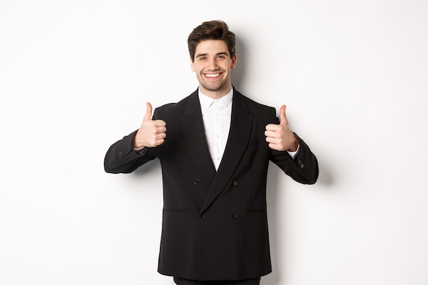 Concepto de fiesta de año nuevo, celebración y estilo de vida. Imagen de atractivo hombre de negocios feliz en traje formal, mostrando el pulgar hacia arriba y sonriendo, como y aprobar, de pie sobre fondo blanco.