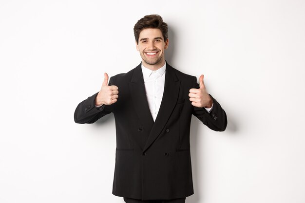 Concepto de fiesta de año nuevo, celebración y estilo de vida. Imagen de atractivo hombre de negocios feliz en traje formal, mostrando el pulgar hacia arriba y sonriendo, como y aprobar, de pie sobre fondo blanco.