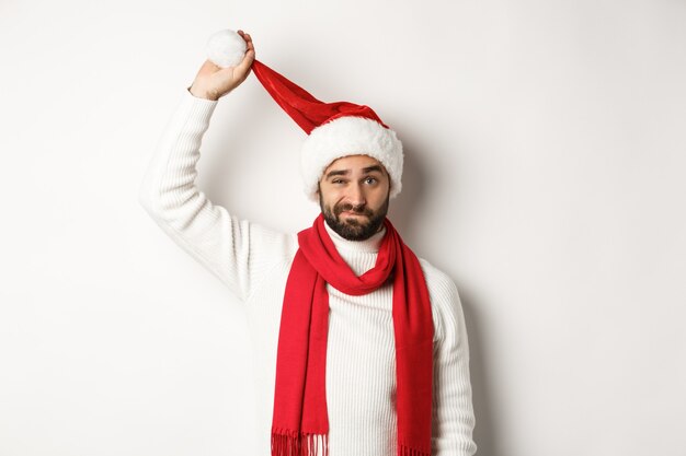 Concepto de fiesta y celebración de Navidad. Chico triste despegando el gorro de Papá Noel y haciendo muecas, mirando molesto, de pie contra el fondo blanco, mal año nuevo