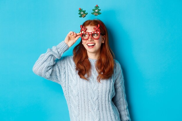 Concepto de fiesta y celebración de Navidad. Linda chica adolescente pelirroja celebrando el Año Nuevo, vistiendo diadema de árbol de Navidad y gafas divertidas, mirando a la izquierda divertido, fondo azul