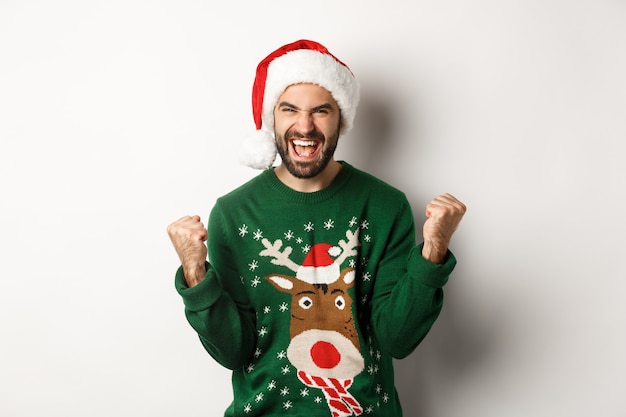 Concepto de fiesta, celebración y vacaciones de Navidad. Chico feliz con gorro de Papá Noel y suéter, haciendo puños y regocijándose, triunfando, de pie sobre fondo blanco.