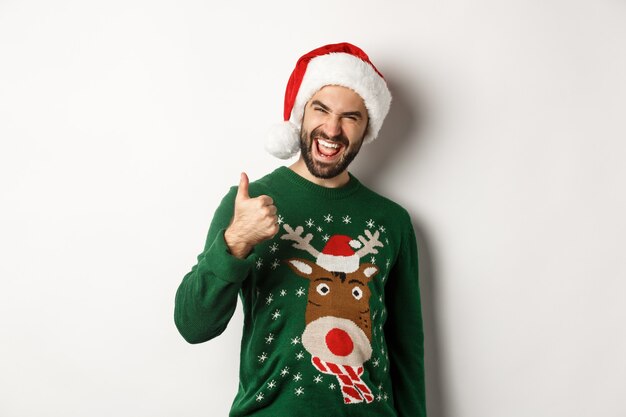 Concepto de fiesta, celebración y vacaciones de Navidad. Hombre disfrutando de año nuevo, mostrando el pulgar hacia arriba en señal de aprobación, vistiendo gorro de Papá Noel, fondo blanco.